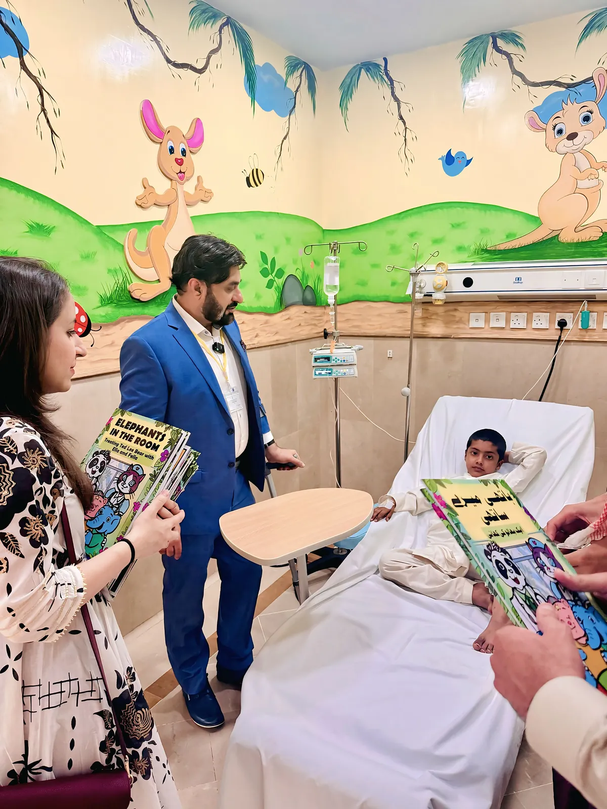 Dr Khan showing a copy of Elephants in the Room to a young reader at their bedside, beneath the ward's painted mural.